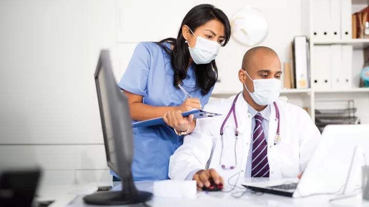 A medium-dark skin toned man in a lab coat uses a laptop. A medium-light skin toned woman in blue scrubs looks over his shoulder, taking notes. Both wear surgical masks. 