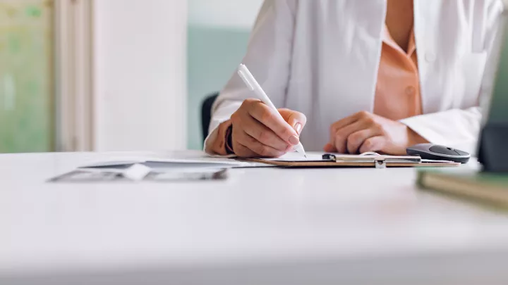 A view of the hands of a woman in a lab coat writing on a medical chart. 