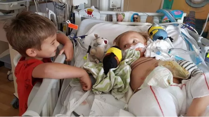 A toddler stands beside a hospital bed, smiling at a baby who is lying down surrounded by stuffed animals and toys.