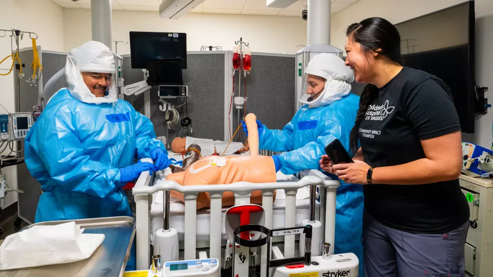 Trainees in full HAZMAT suits work on medical mannequins in a simulated hospital room under the supervision on an instructor
