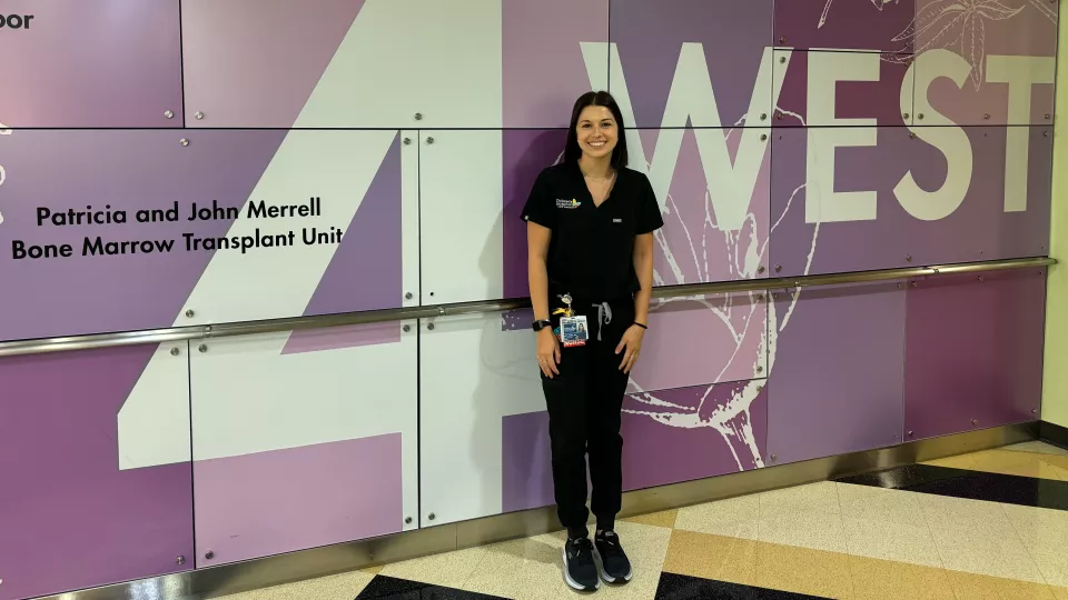 Kate, wearing black scrubs, stands smiling in front of the purple 4 West lobby wall,.