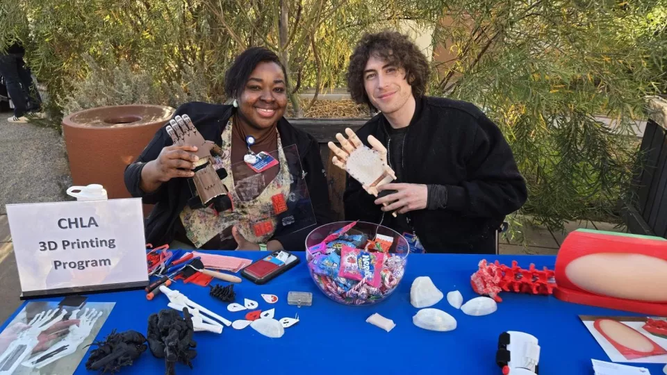 An adult with medium dark skin tone and an adult with light skin tone hold 3D printed prosthetic hands and sit outdoors behind a table displaying 3D-printed medical devices.