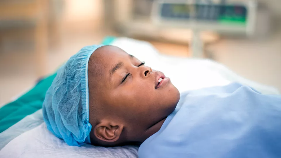 Young child laying on a hospital bed wearing a blue hospital gown and head cap.