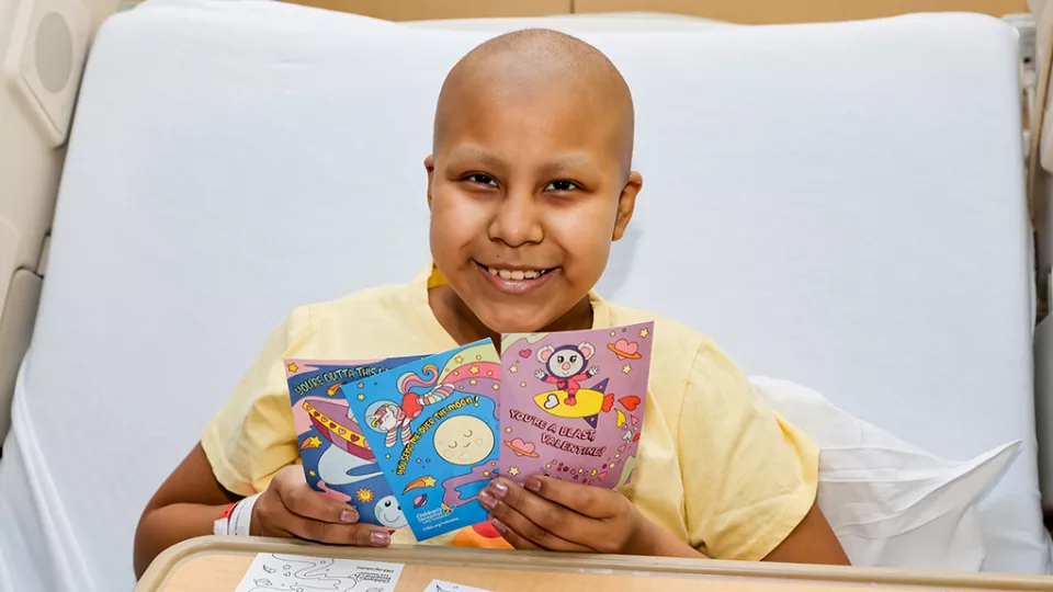 Young patient smiles in their hospital bed as they hold Valentine's Day cards