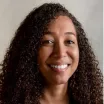 Headshot of a smiling woman with dark skin tone and dark curly hair against a neutral indoor background