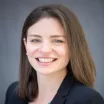 Headshot of a smiling woman with light skin tone and shoulder-length brown hair wearing a dark suit jacket against a neutral indoor background