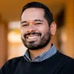 Headshot of a smiling man with medium skin tone, dark hair and a closely cropped beard, wearing a checkered blue dress shirt under a dark sweater against a warm, indoor background