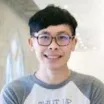 Headshot of a smiling man with light skin tone, short dark hair and glasses wearing a white and grey long-sleeved t-shirt against a brightly lit indoor background