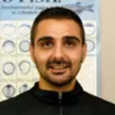 Headshot of a smiling man with medium skin tone, dark hair and close cropped beard wearing a zipped turtleneck against an indoor background