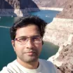 Selfie of a man with medium skin tone and dark hair wearing browline eyeglasses and a light colored t-shirt against the background of the exposed sharp cliffs of a deep reservoir