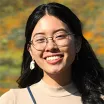 Headshot of smiling woman with light skin tone and long dark hair wearing eyeglasses and a tan turtleneck against a blurred outdoor background