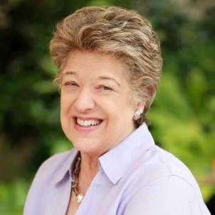 Smiling woman with light skin tone and light brown hair against background of blurred green foliage