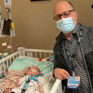 Male infant with light skin tone lies on their back in hospital crib as adult male with light skin tone and shaved head wearing glasses and a surgical mask leans on the crib's rail