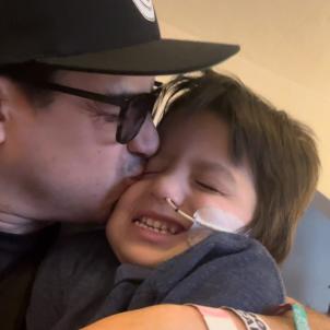 Father with medium skin tone wearing black cap and sunglasses kisses smiling young son on the cheek