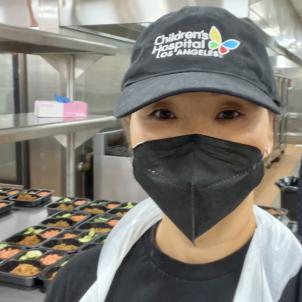 A medium-light skin toned woman in a KN-95 mask, Children's Hospital Los Angeles cap and plastic apron stands in a kitchen.