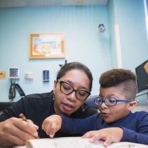 A woman holds a pencil to a book while a young boy with glasses points to the page.