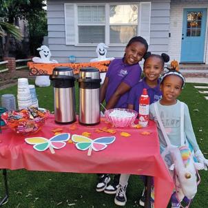 Three young girls with dark skin tone smile as they sell hot chocolate on Halloween