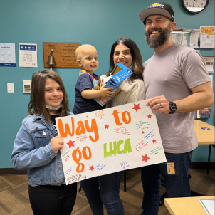 Young girl, toddler, adult woman and adult man stand in front of bell holding sign