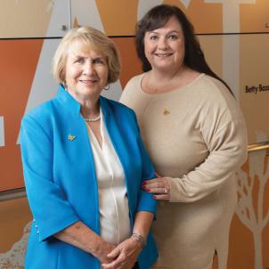 A woman with light skin tone, short blonde hair wearing a light blue jacket standing next to a woman with long dark hair wearing a beige dress, in a hospital corridor.