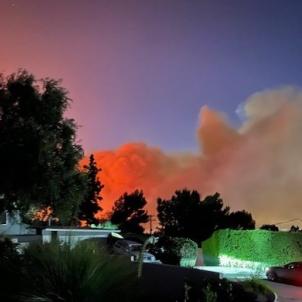The driveway of a home with orange and grey smoke rising above trees along a dark horizon with a purple sky.