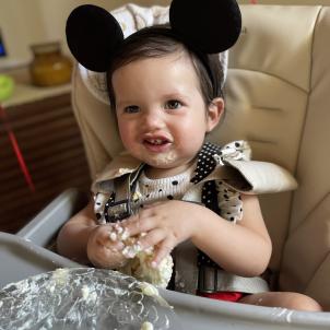 A one year old wearing mickey ears sits in a high chair eating birthday cake.