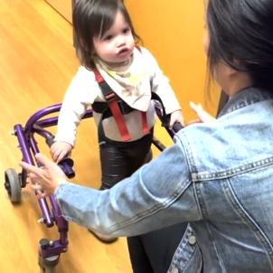 A toddler walks toward her physical therapist using a walker.