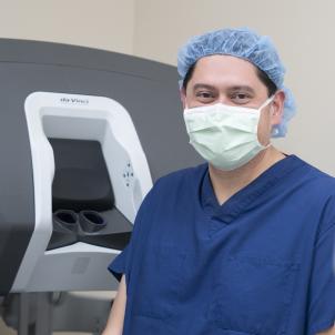 Close-up shot of a man with medium skin tone in dark blue scrubs and surgical mask and cap in front of a large console