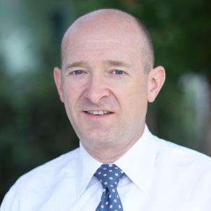 Headshot of a smiling man with light skin tone and shaved head wearing a white dress shirt and blue tie against a blurred outdoor background