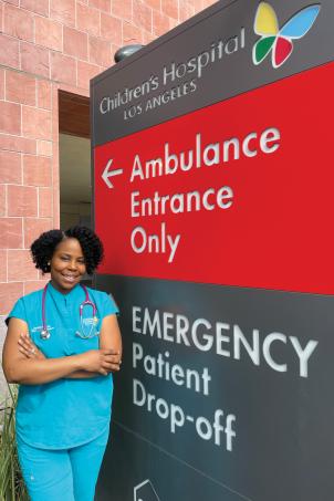 Female doctor with dark skin tone and short dark hair smiles as she stands with her arms crossed outside Children's Hospital Emergency Department entrance