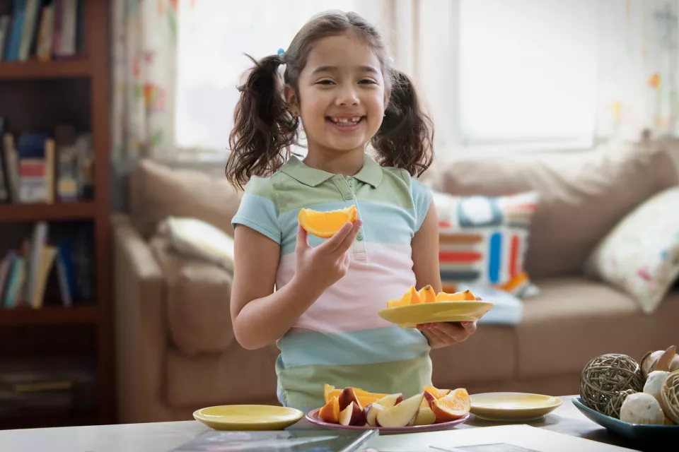 A girl with medium-light skin tone holds a plate of fruit and smiles. She has pigtails and a missing front tooth.