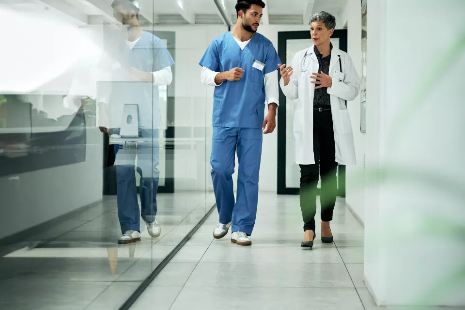 A medium-light skin toned woman with short gray hair wearing a lab coat and stethoscope talks to a medium skin toned man in blue scrubs as they walk. 