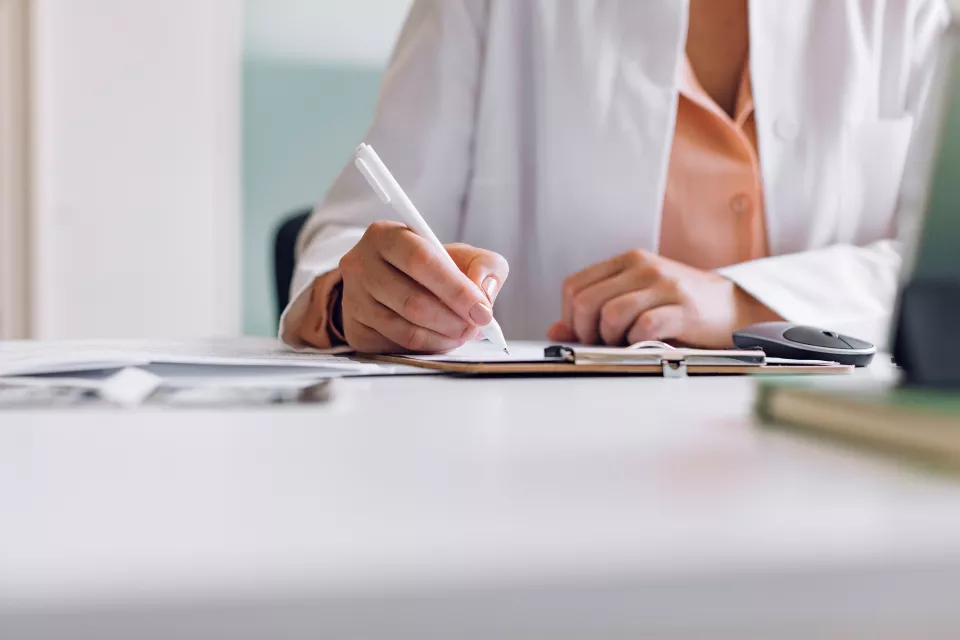 A view of the hands of a woman in a lab coat writing on a medical chart. 