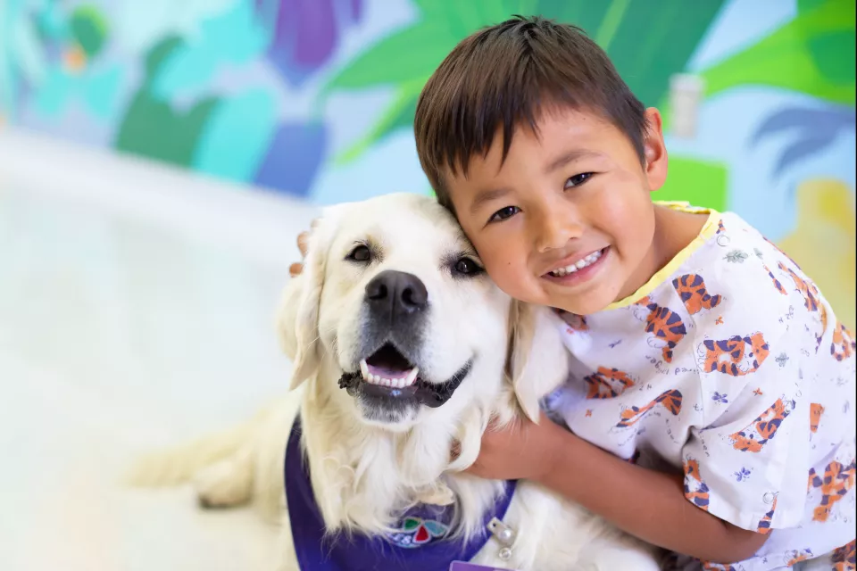 Young boy with medium skin tone and dark hair wearing a tiger print hospital gown smiles as he hugs a Labrador retriever with light colored fur