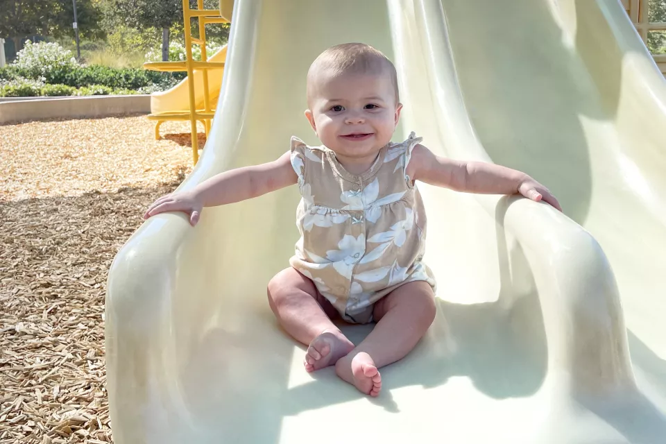 Toddler with medium skin tone smiles as they sit at the bottom of a playground slide