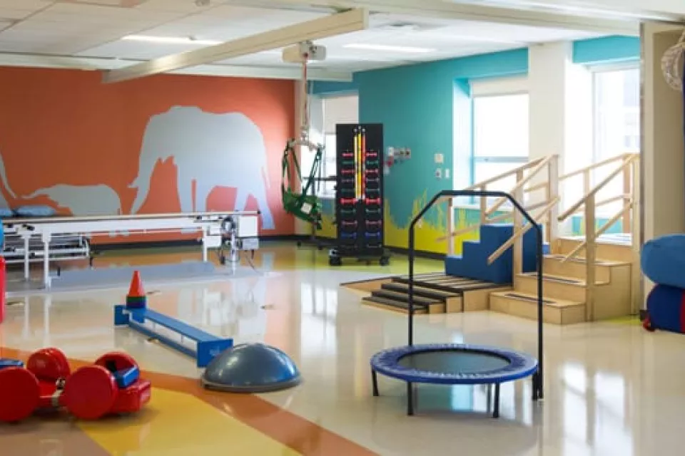 Room with bright colored walls, small trampoline, balance beam and other rehab equipment.